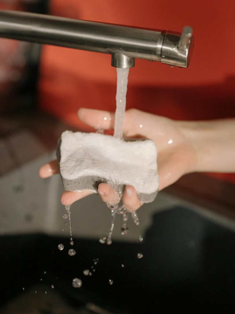 Close-up of two hands rinsing a white sponge under a running faucet, with water droplets falling into a sink, against a blurred red background